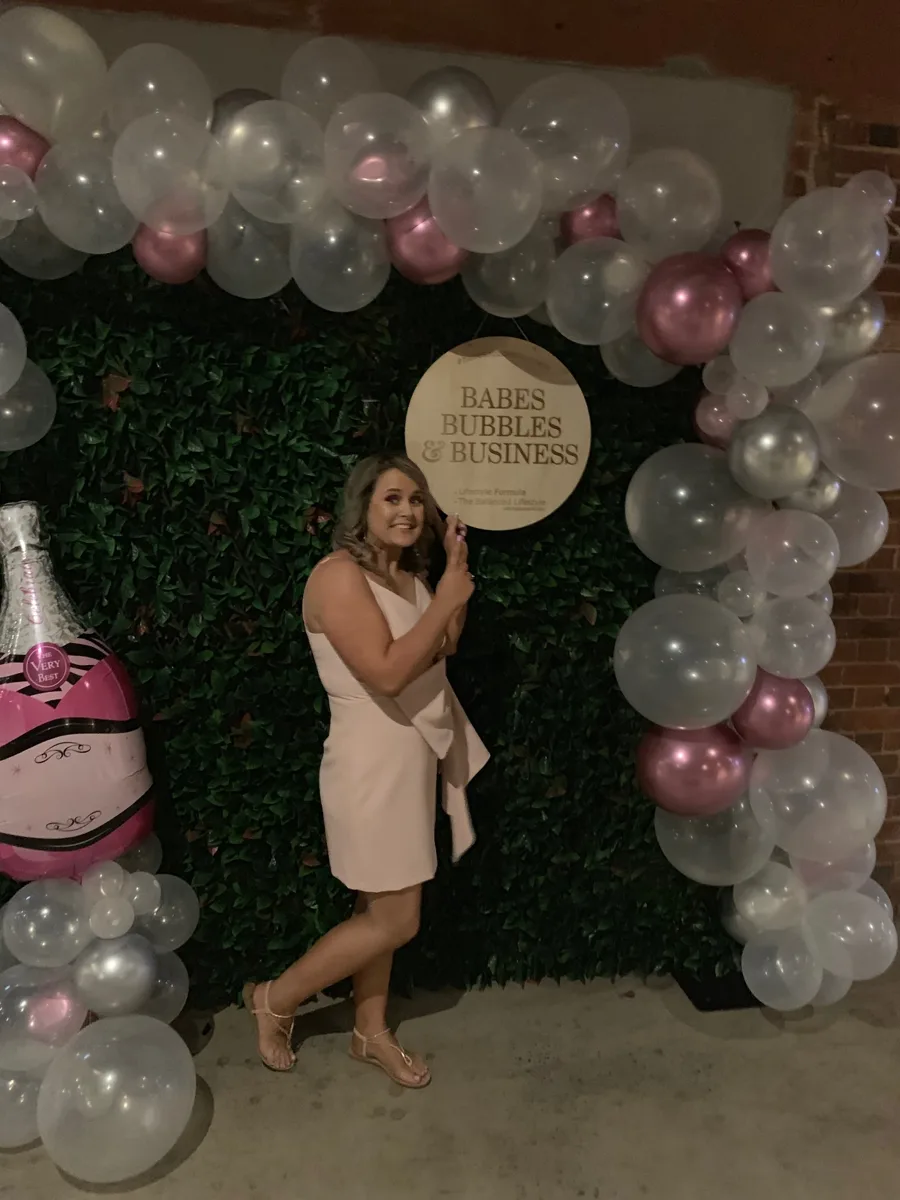 Beck Smith at a Babes Bubbles and Business event backdrop with balloons and a hedge wall