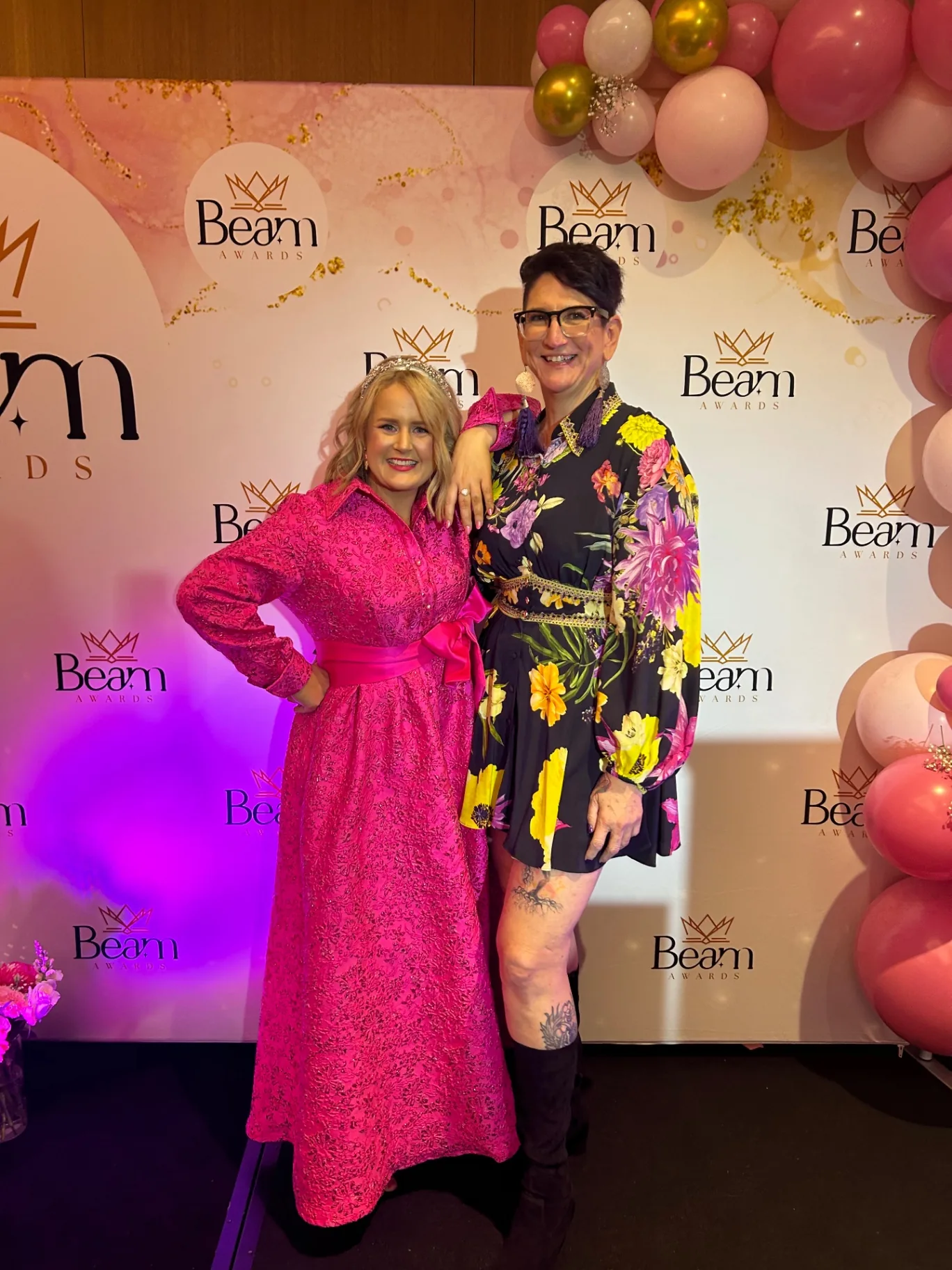 Beck Smith and a guest at the Beam Awards backdrop with pink and gold balloons