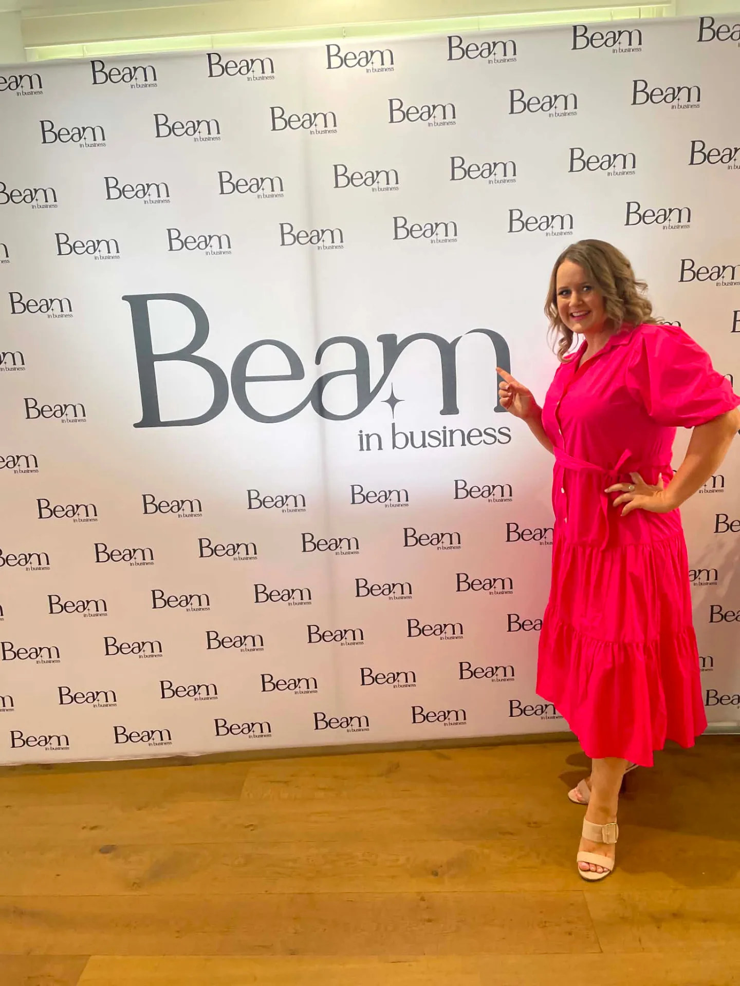 Beck Smith in a pink dress pointing to the Beam in Business logo on the launch media wall